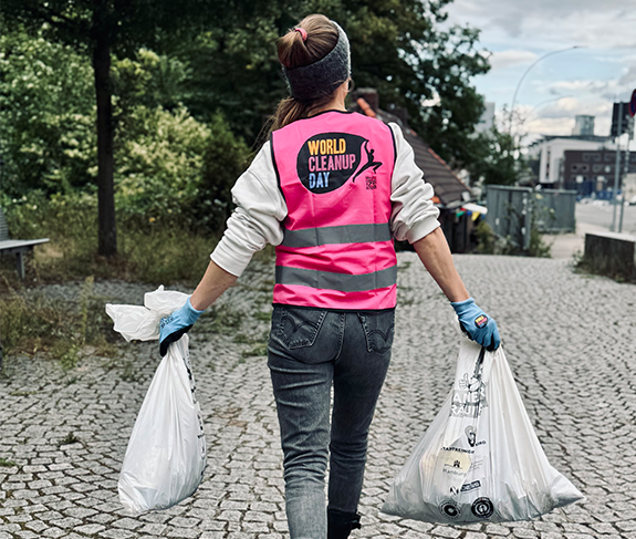 Foto von Verena Rateike auf dem sie mit jeweils einem Müllsack in jeder Hand von der Kamera weggeht. Sie trägt eine pinke Warnweste mit der Aufschrift "World Clean Up Day"
