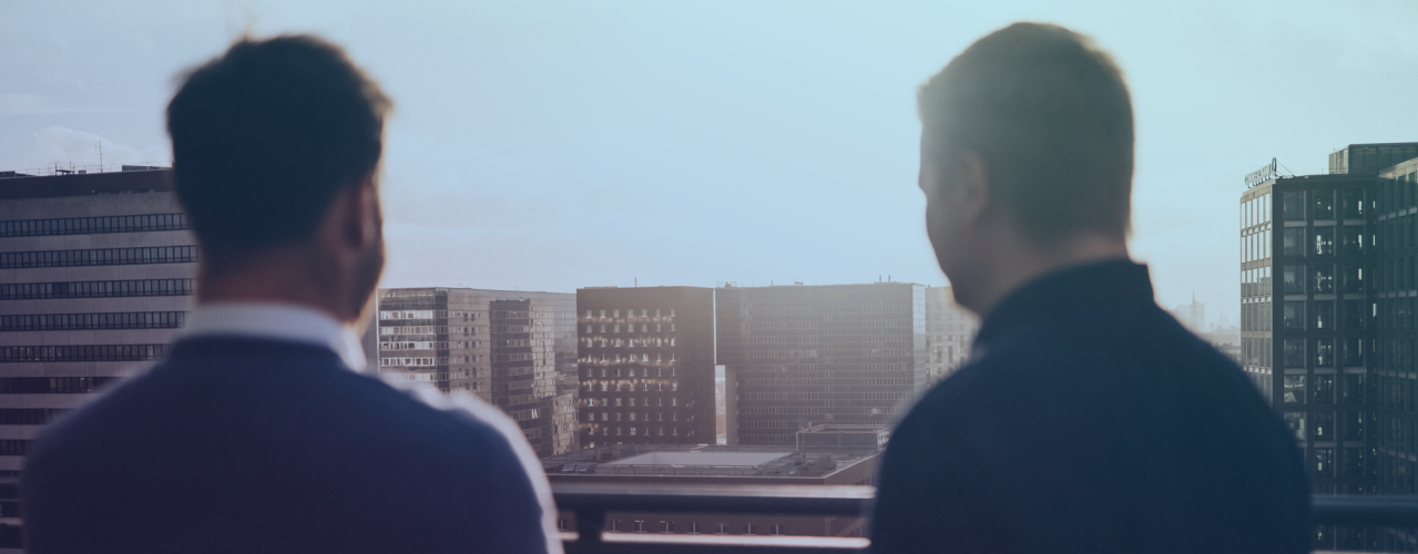 Oliver Haus und Sascha Pohle stehen mit dem Rücken zum Betrachter auf einem Balkon. Vor ihnen geht die Sonne über den Häusern der Stadt unter.