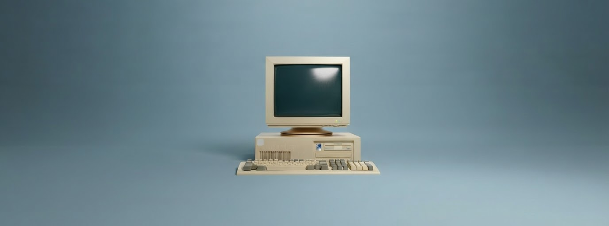 Retro-Desktopcomputer mit Röhrenmonitor, Rechner und Tastatur vor hellblauem Hintergrund. Der beige Computer im Stil der 1980er- und 1990er-Jahre steht frontal im Bild, der Bildschirm ist ausgeschaltet. 