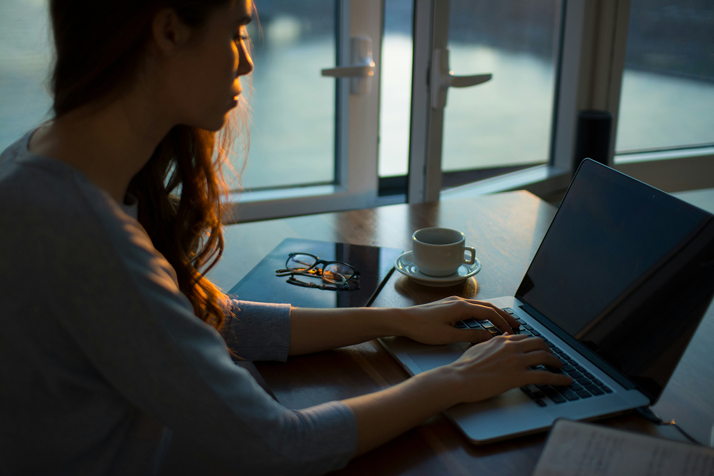 Eine Frau sitzt an einem offenem Fenster an ihrem Laptop. Neben ihr steht eine weiße Kaffeetasse und eine Brille.
